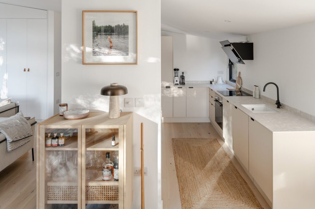 A light, modern kitchen with pale cabinetry, integrated appliances, and a long woven jute runner, shown alongside a cosy living corner featuring a wooden cabinet with glassware, a sculptural table lamp, and framed artwork on a white wall. Soft natural light creates gentle shadows across the space.