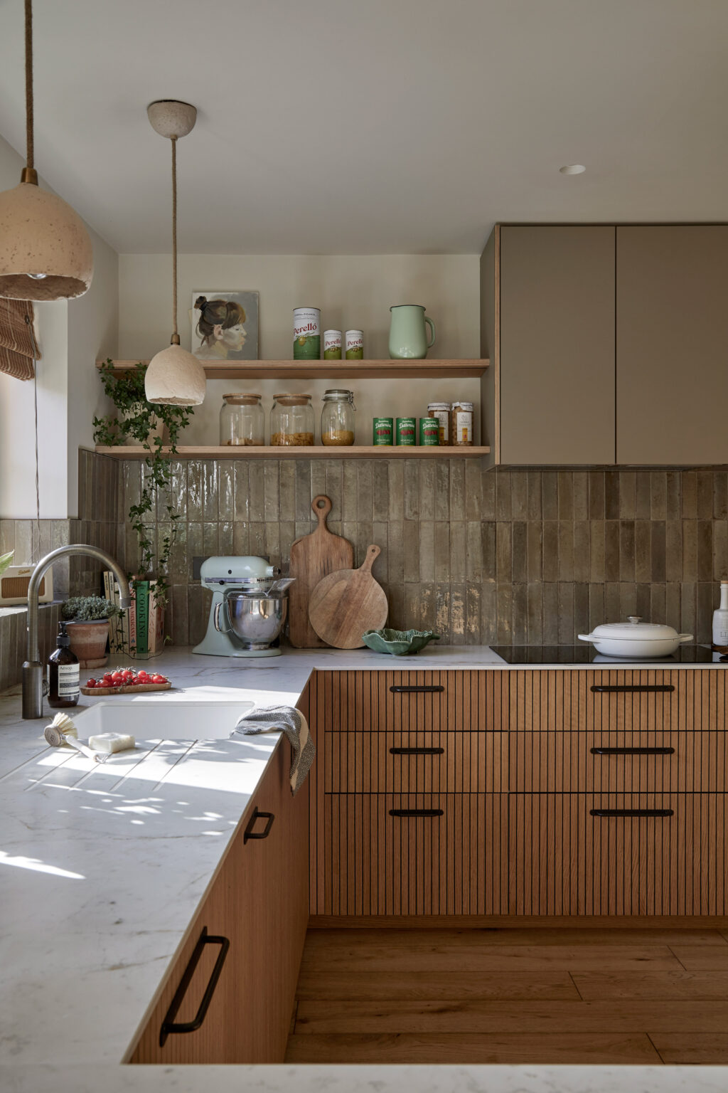 A calm and cosy kitchen with a natural colour palette, featuring rustic green tiles, natural wood details and soft stone-effect worktops. Bespoke open shelving displays a trailing plant, painted canvas and decorative ambient items.