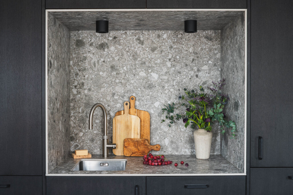 A built-in niche with a stone backsplash holds several wooden cutting boards beside a small sink with a chrome tap and red grapes on the countertop