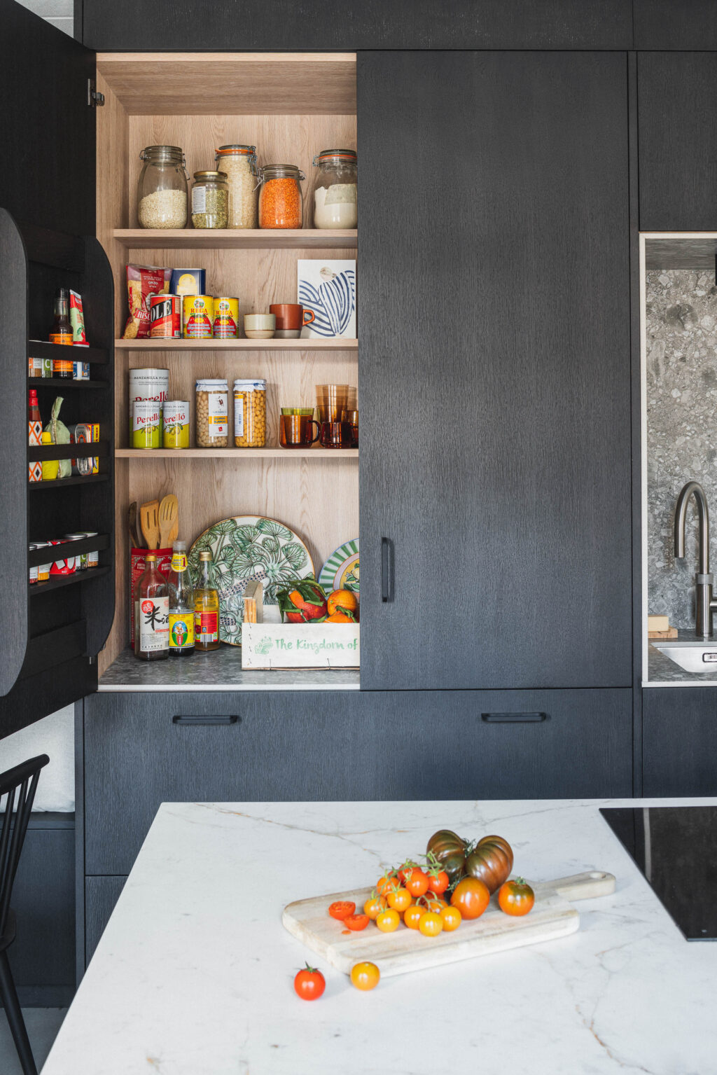 An open pantry with food in jars on wooden shelves. A white kitchen island is in front with tomatoes on it.