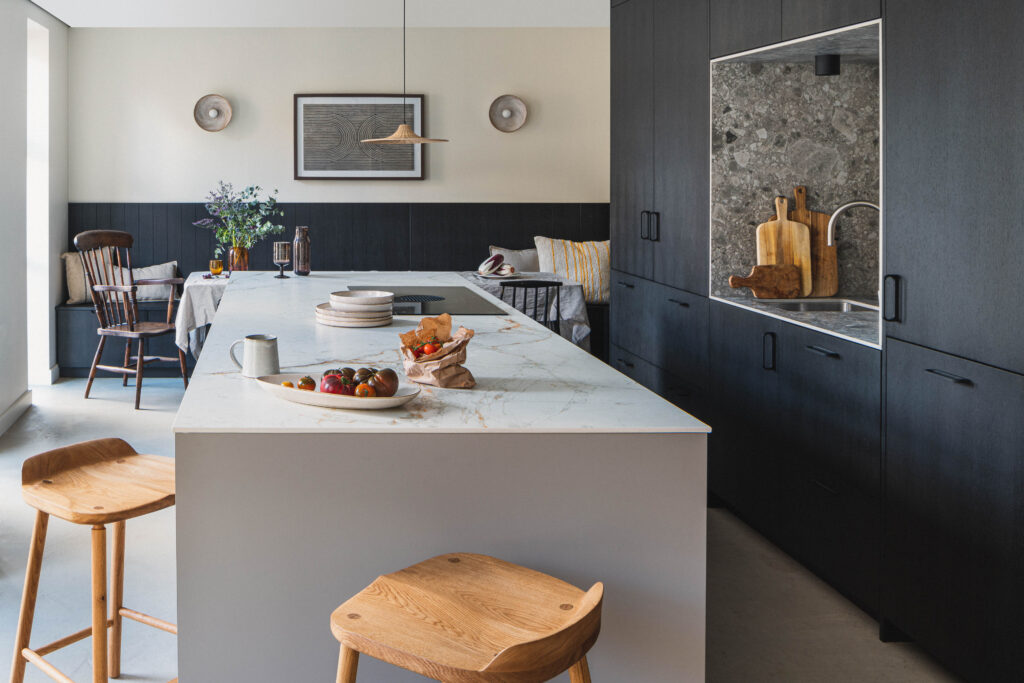 A modern kitchen with a large white marble island, two wooden bar stools, and dark wood cabinetry. A built-in niche with a stone backsplash holds several wooden cutting boards beside a small sink. In the background, a cosy dining nook with dark paneling features a round table, wooden chairs, cushions, and decorative wall art. Fresh produce and tableware are arranged neatly on the island.