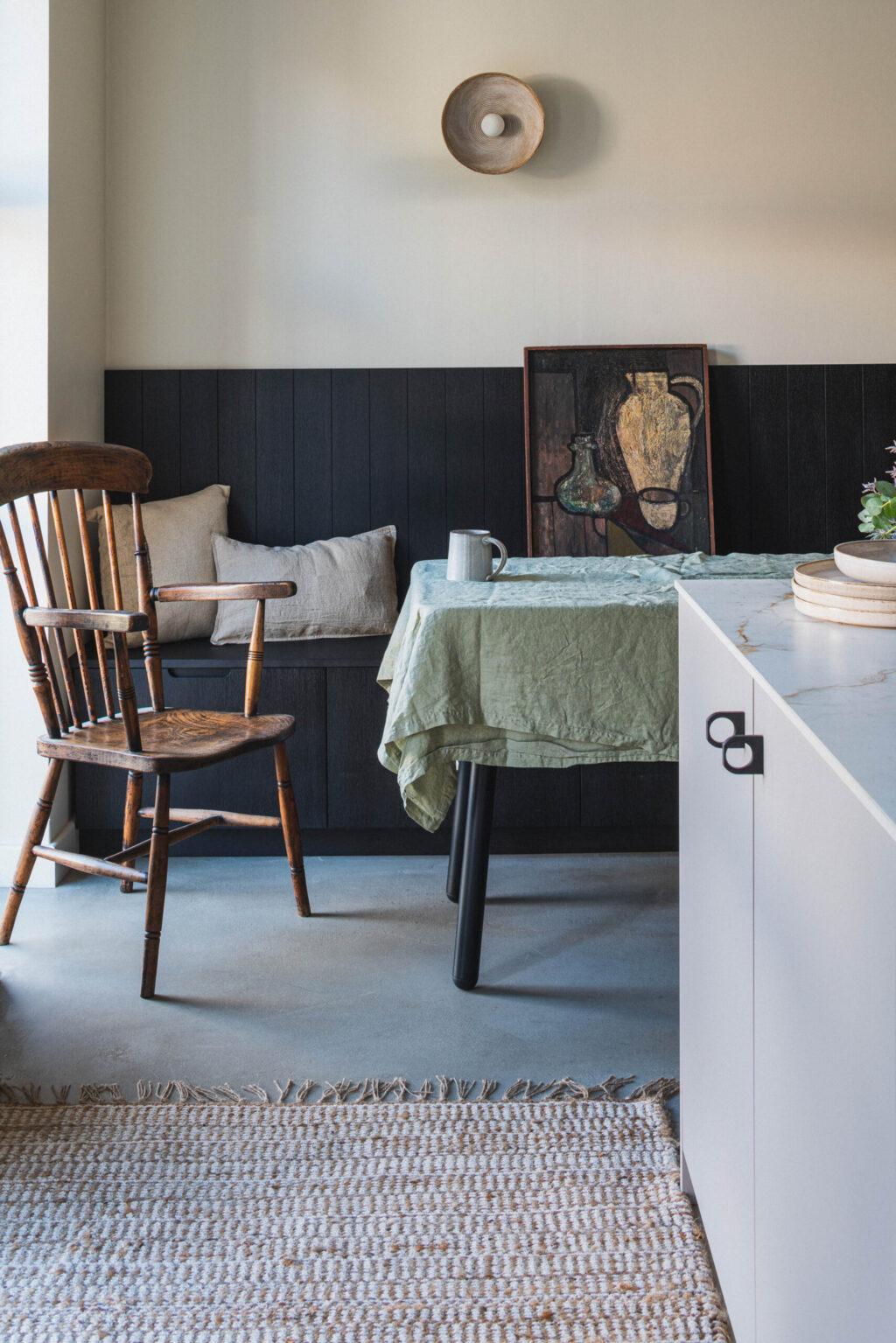 A cozy dining nook with a black wood-paneled bench, beige cushions, and a wooden chair beside a window. A table covered with a wrinkled green cloth holds simple ceramic dishes. A framed still-life painting of vases rests against the wall above the bench, and a round wall sconce hangs above it. The scene has a warm, rustic, and lived-in feel.