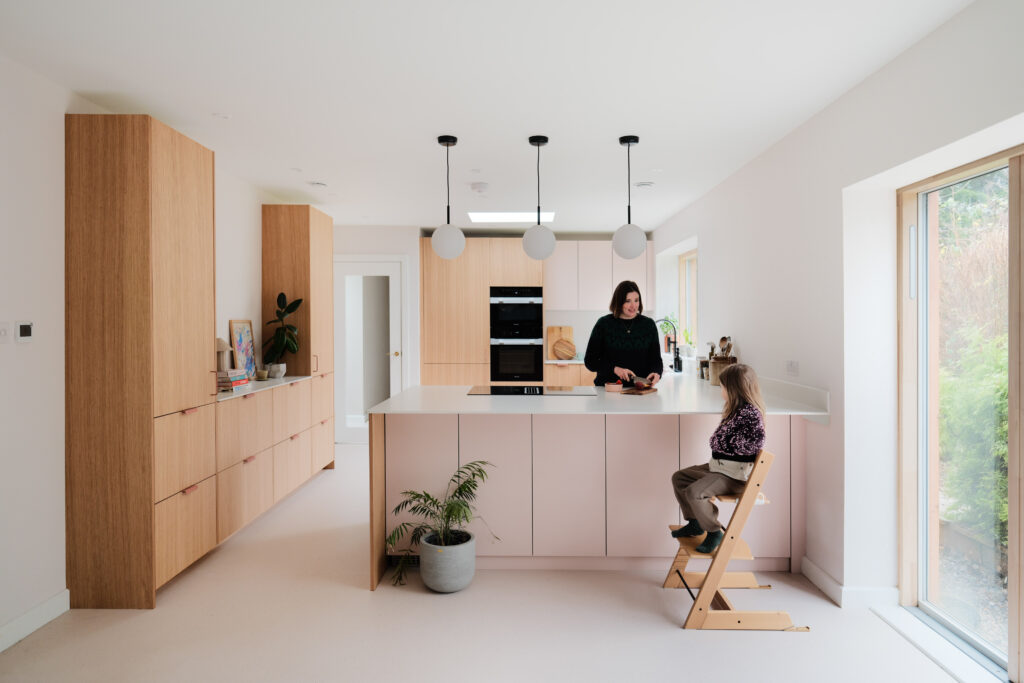 A modern, light-filled kitchen with pale pink lower cabinets, wood-paneled tall cabinets, a women is chopping food for a child who is on a chair infront on the kitchen island. A large white island with an induction cooktop, and three round pendant lights hanging above. The countertops are white, and a potted plant sits at the base of the island. The space features clean lines, minimal decor, and natural light from a window on the right.