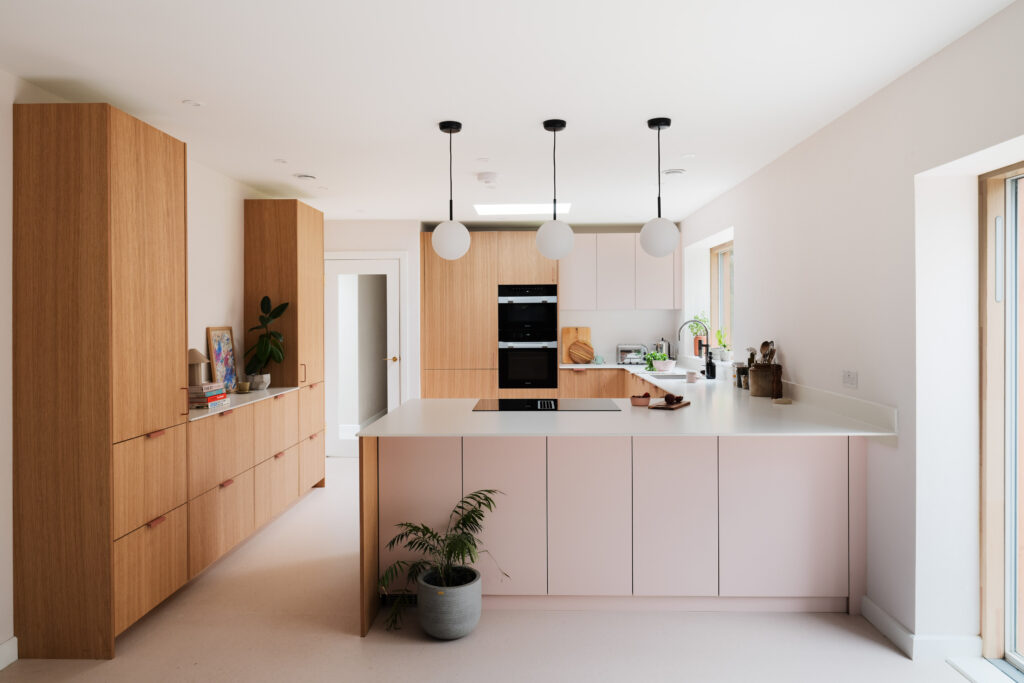 A modern, light-filled kitchen with pale pink lower cabinets, wood-panelled tall cabinets, a large white island with an induction hob, and three round pendant lights hanging above. The countertops are white, and a potted plant sits at the base of the island. The space features clean lines, minimal decor, and natural light from a window on the right.