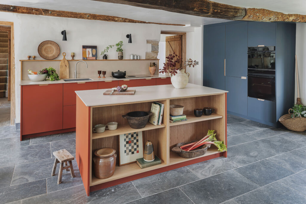 Rustic modern kitchen with a central island featuring open wooden shelving and a pale countertop, terracotta-red cabinetry, exposed wooden beam, farmhouse-style wooden doors, stone floor, blue kitchen cabinetry on the side of the kitchen with a built in oven in and minimal decor including pottery, plants, and cutting boards on a simple wall shelf.