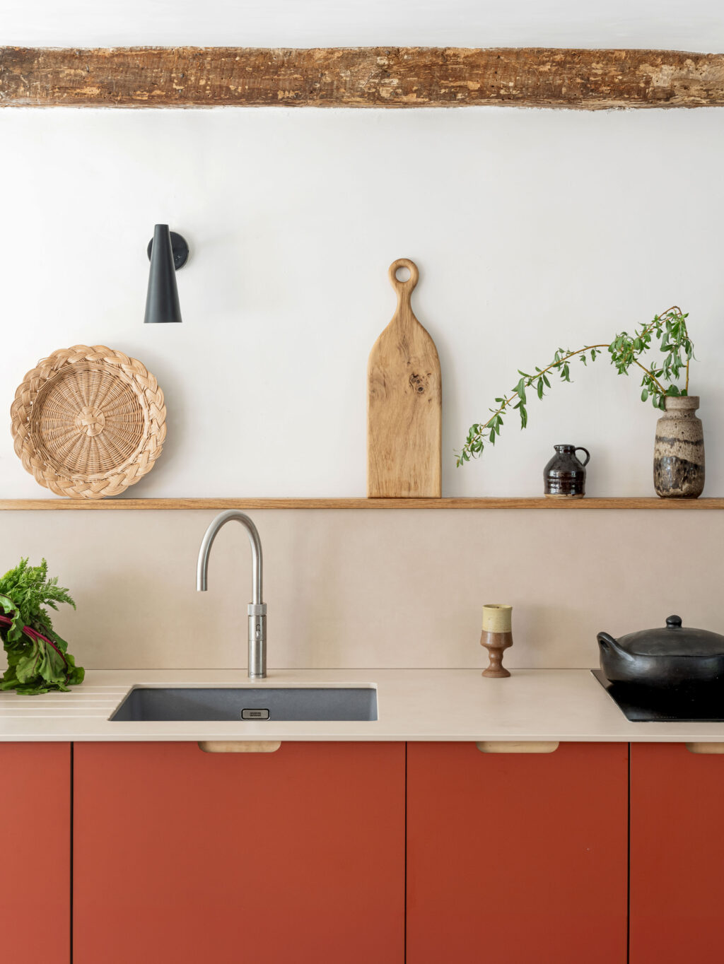 Rustic modern kitchen featuring a pale countertop, terracotta-red cabinetry and minimal decor including pottery, plants, and cutting boards on a simple wall shelf.