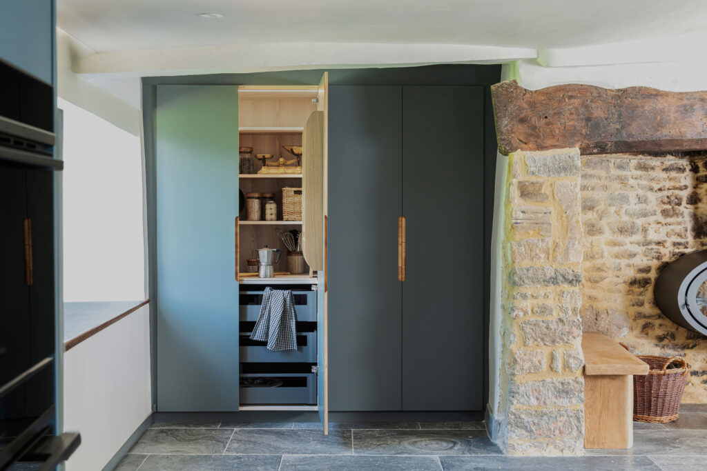 A floor to ceiling blue built in pantry cupboards with kitchen ware on the shelving next to a rustic stone fireplace with stone flooring.