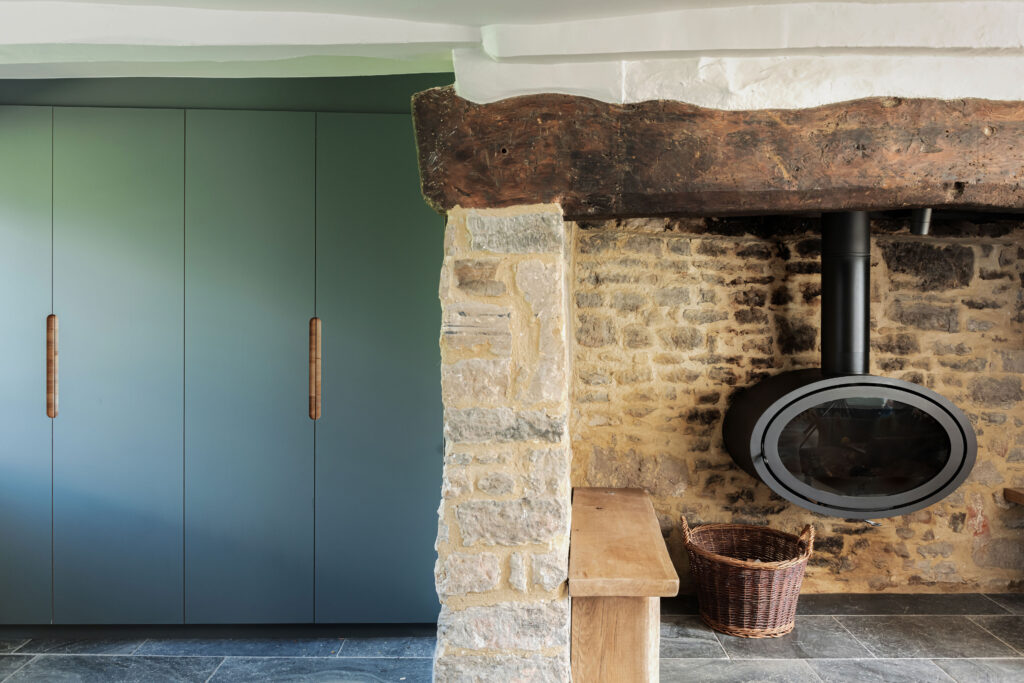 Modern blue tall cabinetry next to a modern log burner in a rustic stone fireplace with minimal, modern styling of wooden benches and a brown arm chair.