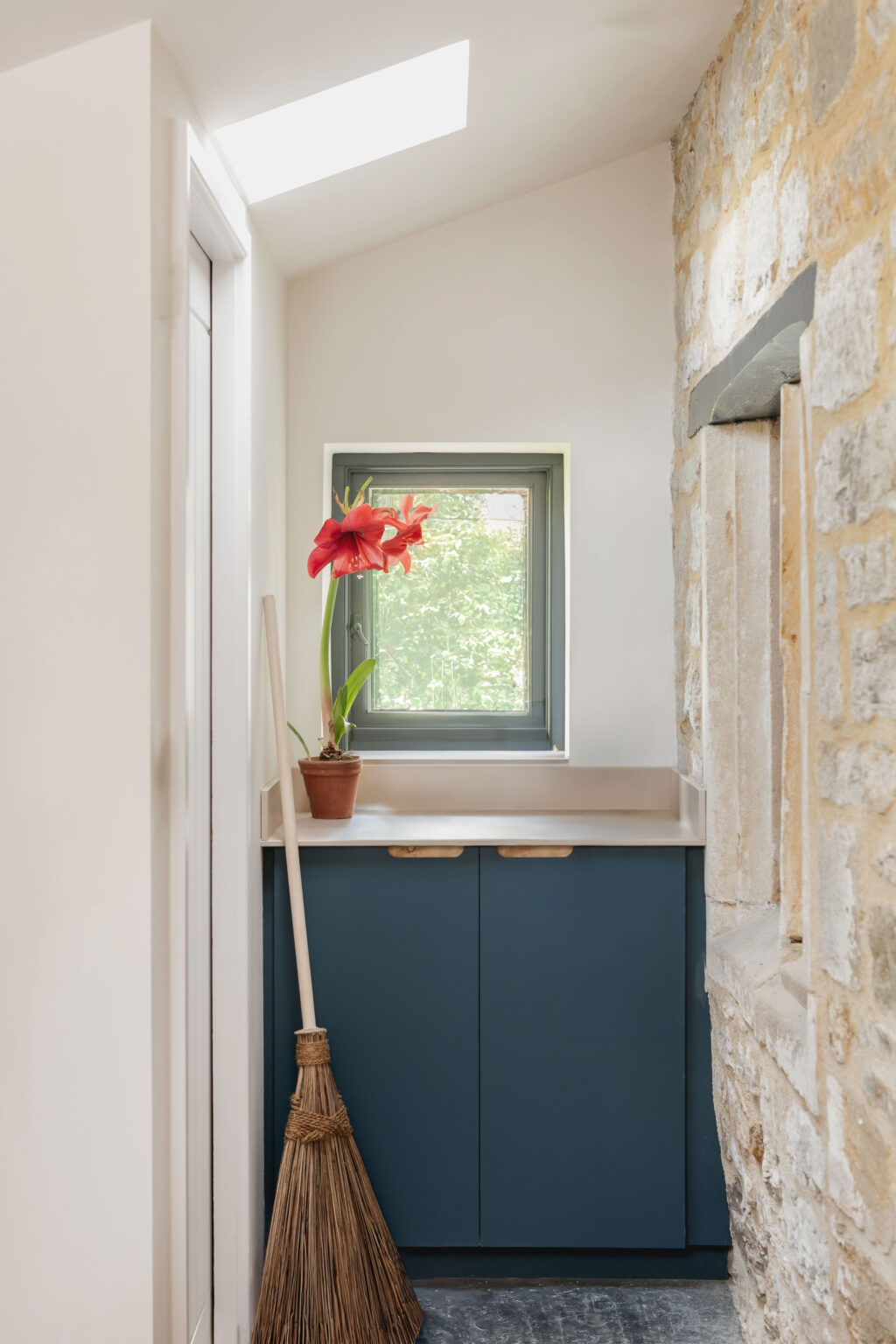 A utility room with a blue cabinet under a small window, a wooden broom in leant against the cabinets with a potted red flower on the counter top.