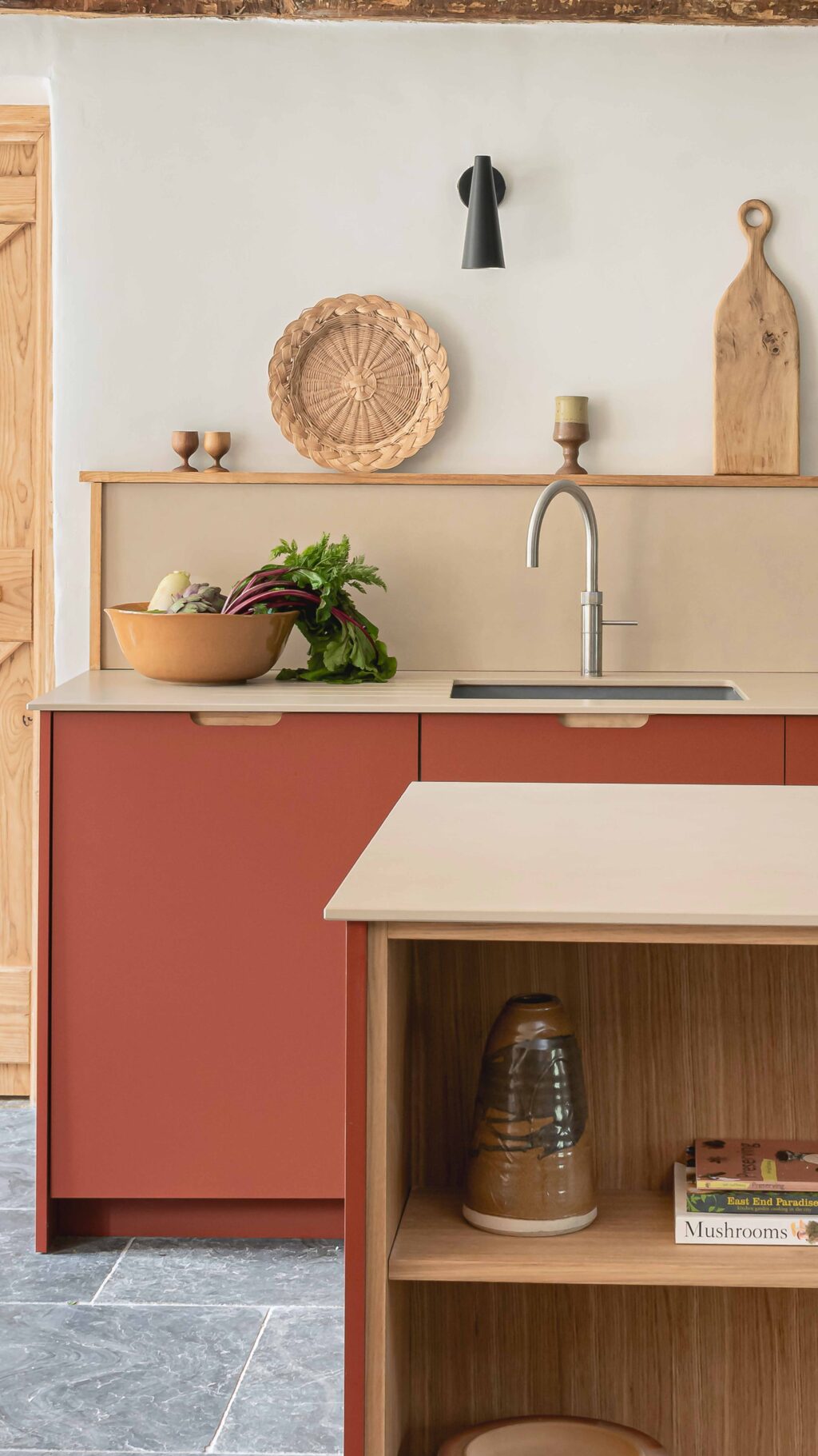 Rustic modern kitchen with a kitchen island on the side featuring open wooden shelving and a pale countertop, terracotta-red cabinetry, farmhouse-style wooden doors, stone floor, and minimal decor including pottery, plants, and cutting boards on a simple wall shelf a bowl is next to the sink filled with vegetavbles.