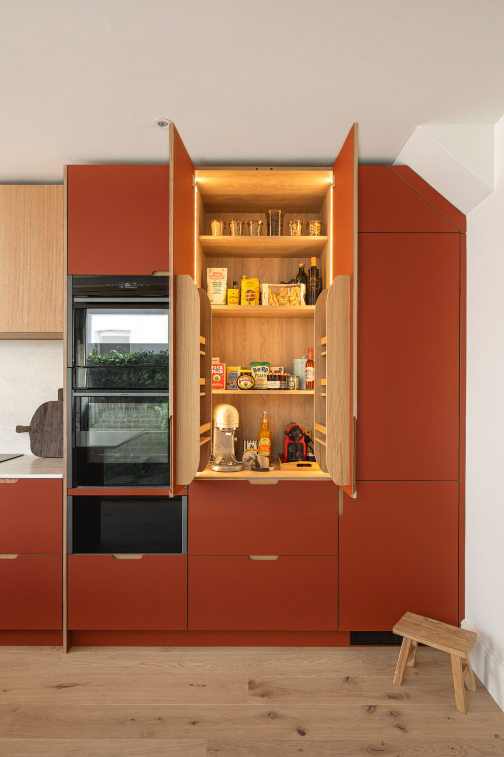 Modern kitchen with matte terracotta cabinetry and light wood accents, showing a tall pantry cabinet with doors open to reveal wooden shelves stocked with food items and drinks beside built-in black ovens
