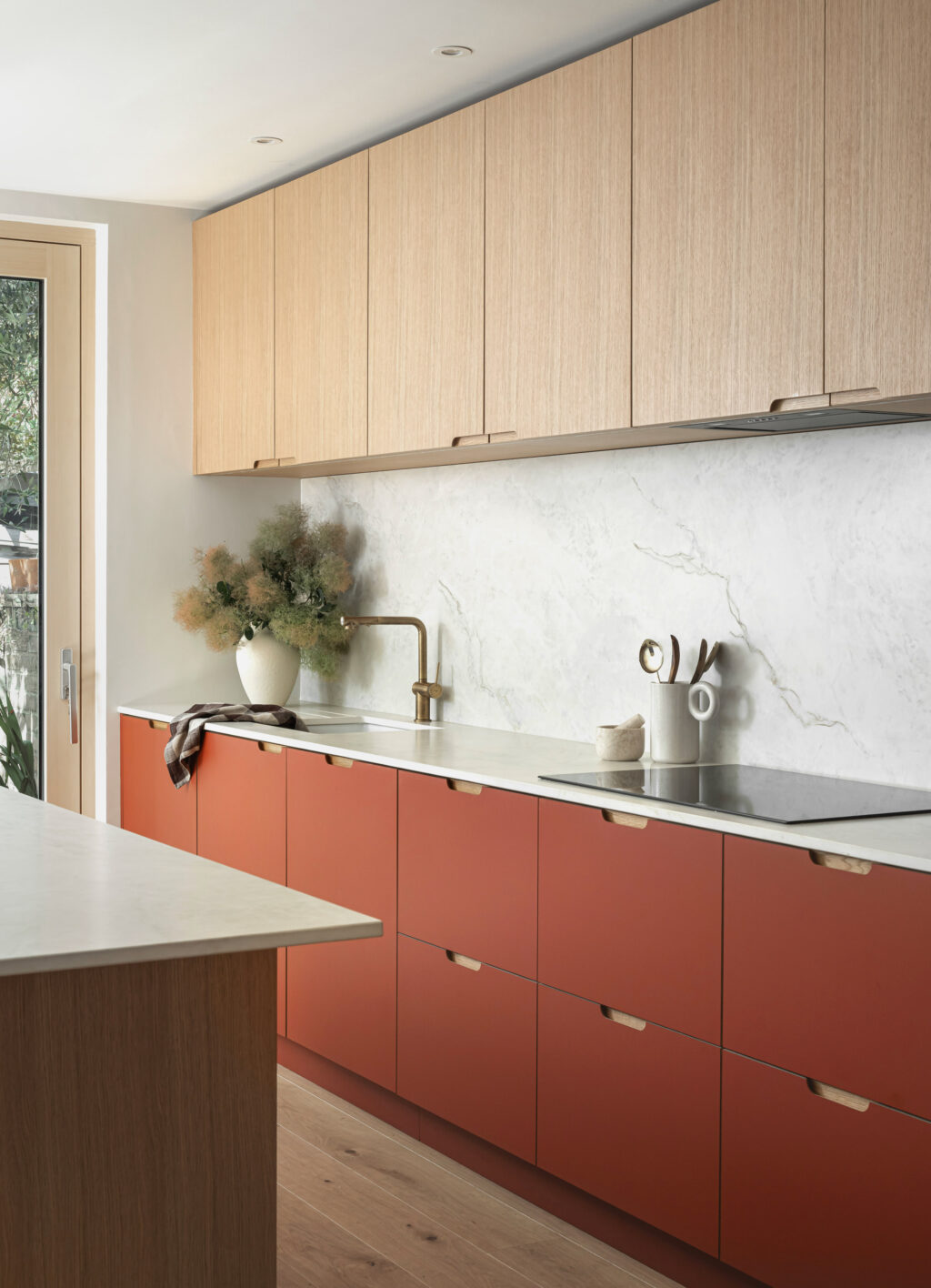Contemporary kitchen with matte terracotta lower cabinets, light wood upper cabinets, white marble backsplash, and built-in black ovens beside a large window, featuring light wood flooring and a minimalist island.