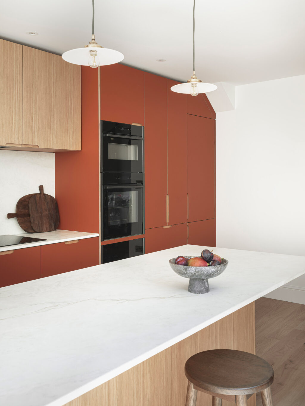 Contemporary kitchen with matte terracotta lower cabinets, light wood upper cabinets, white marble backsplash, and built-in black oven featuring a minimalist island with a fruit bowl.