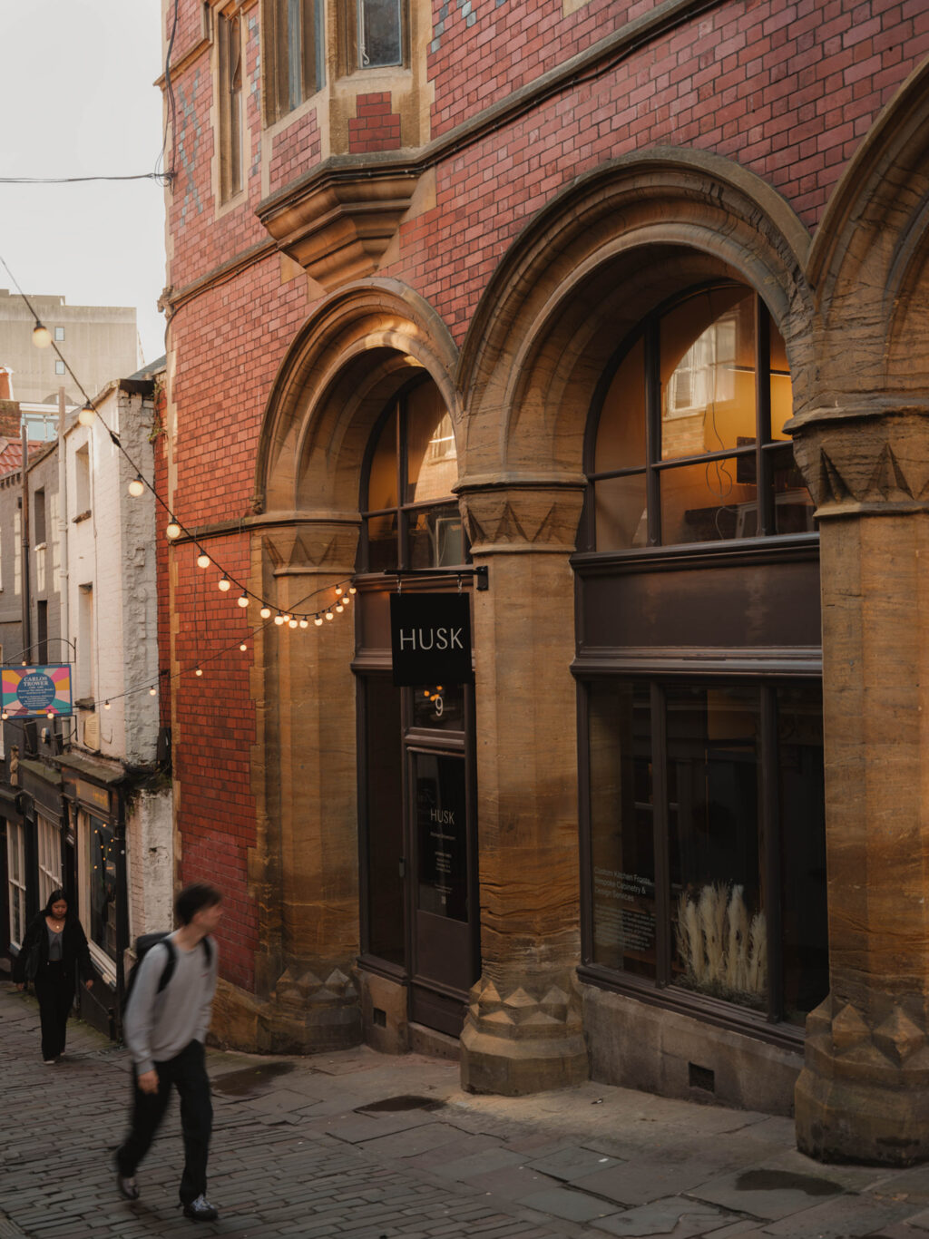 A person walking up a narrow cobblestone street past a shopfront with arched stone architecture. A black hanging sign reads ‘HUSK,’ and string lights are hung above the entrance.