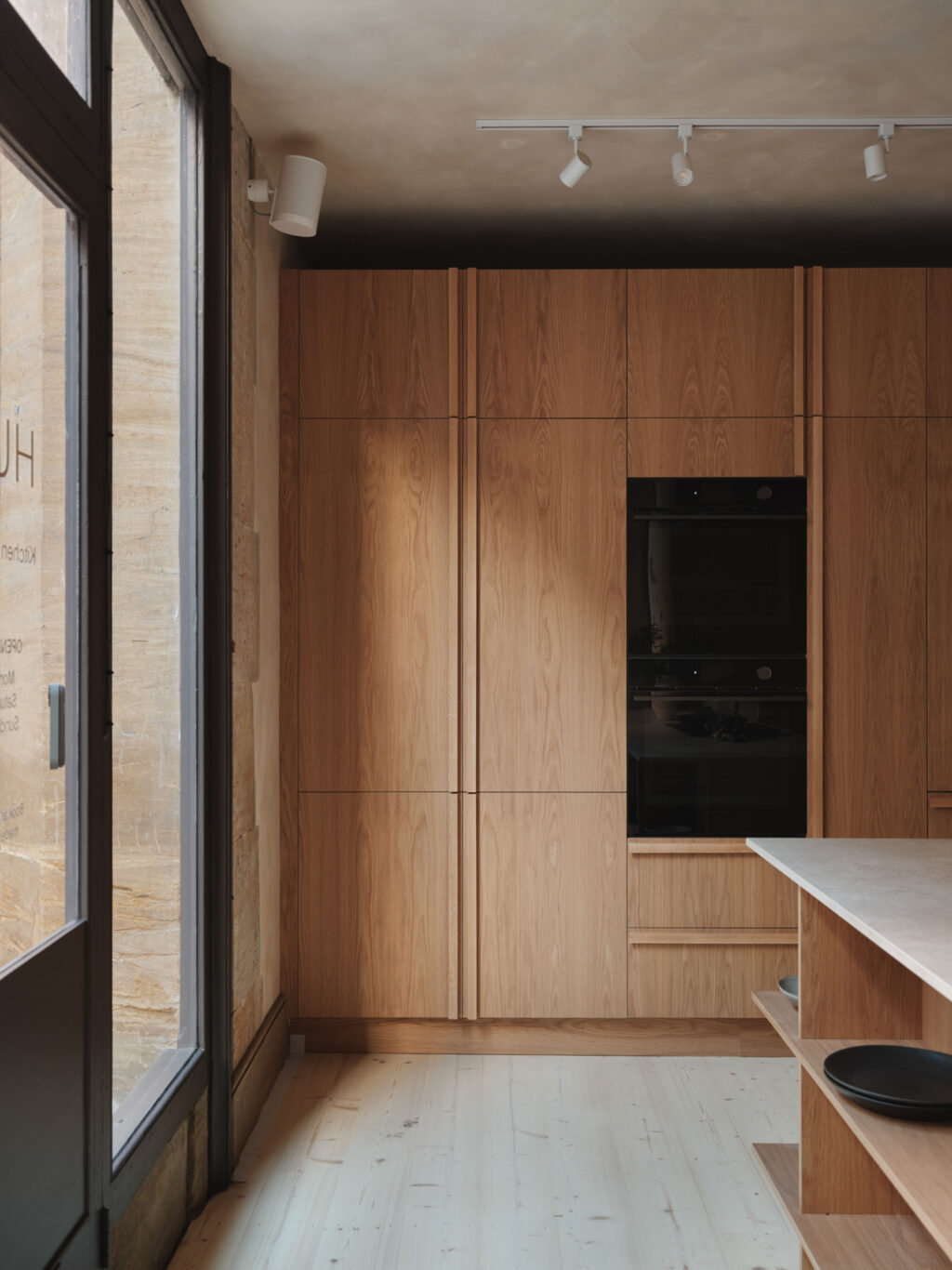 Modern oak kitchen cabinets from floor to ceiling with a build in black oven next to a large window.