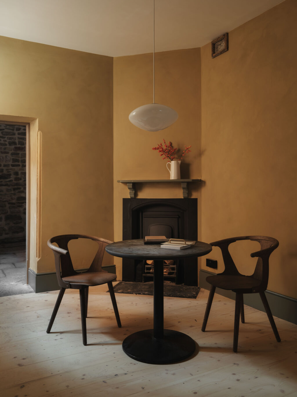 A minimal yellow room with a white pendant light, two dark wooden chair and black round table with a black fireplace behind.