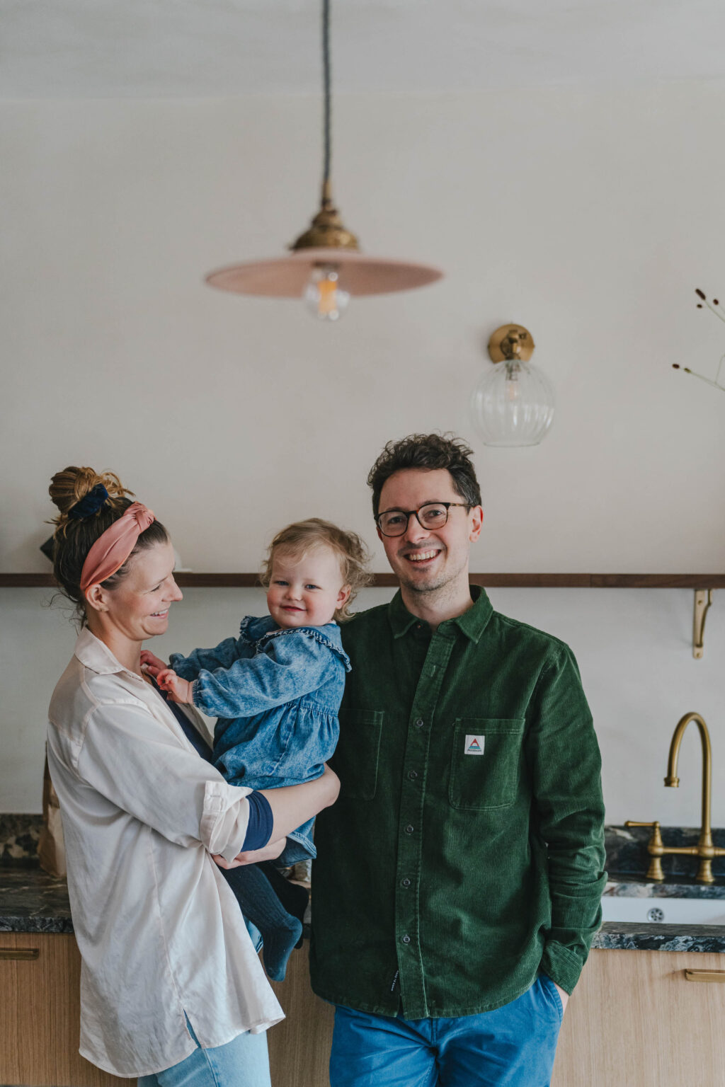 A husband and wife with a toddler in the mothers arms in a modern white and wooden kitchen with black marble countertops