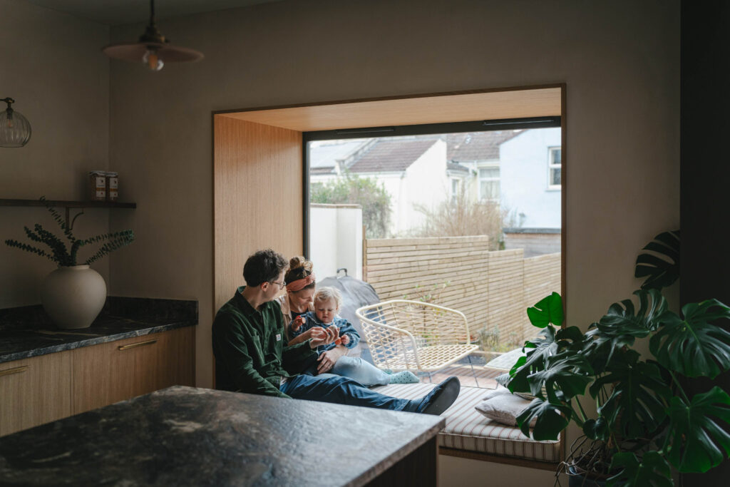 A family of a husband, wife and toddler sat in a window seat playing with the toddler. The window seat is in a modern wooden kitchen with neutral walls with a large green plant in the corner.