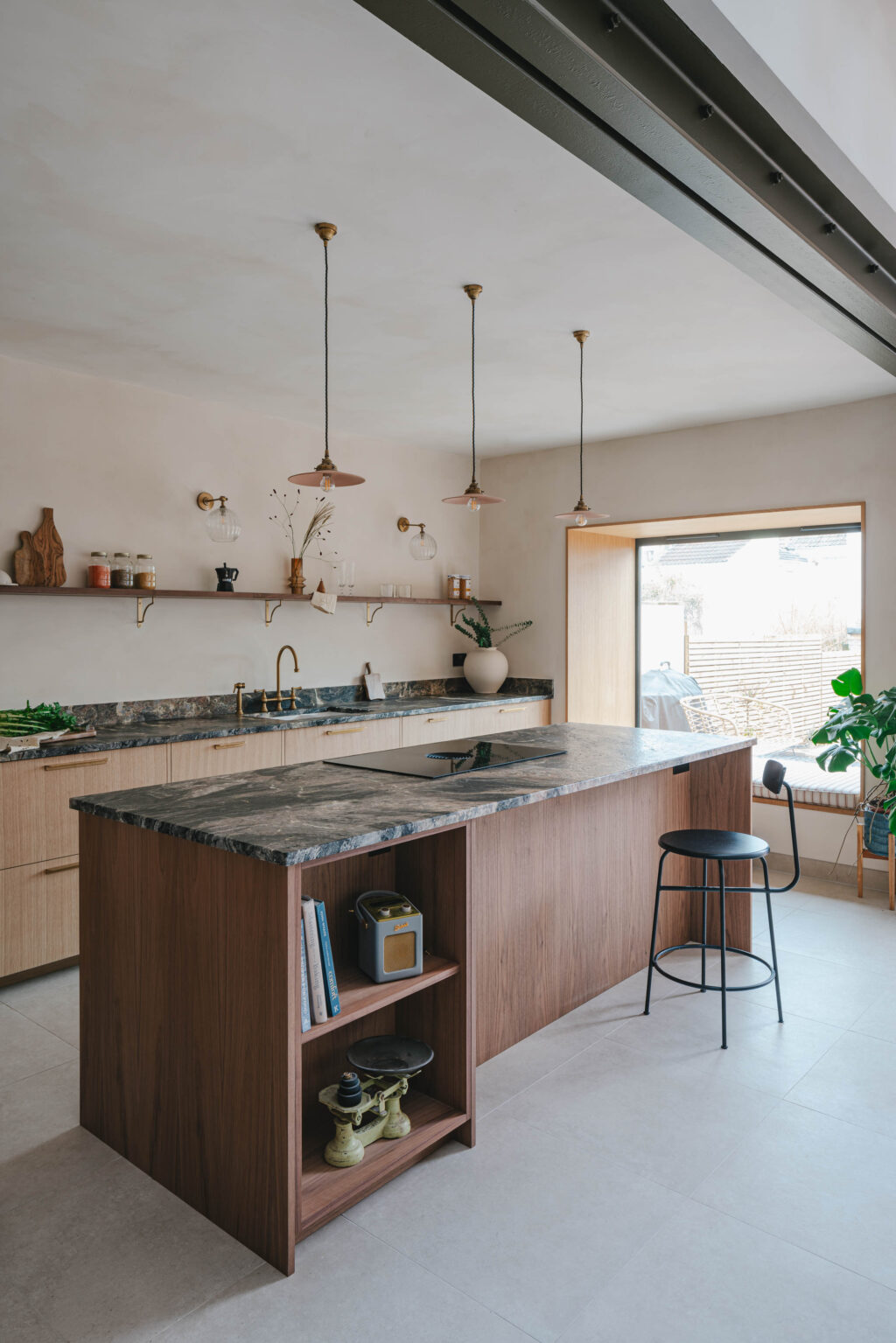 Light-filled modern kitchen with walnut and oak cabinetry, a large island topped with dark stone, open wall shelving with jars and ceramics, brass fixtures, pendant lights, and a window seat beside a sliding glass door overlooking an outdoor space.