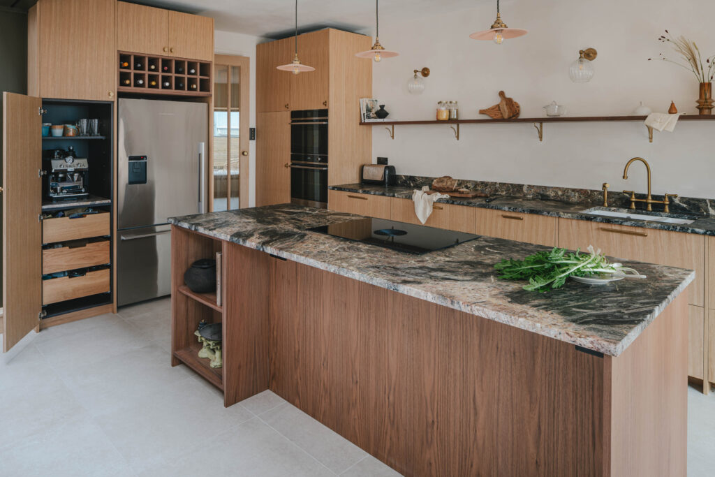 Modern kitchen with walnut and oak cabinetry, a large stone-topped island with an induction cooktop, built-in stainless steel fridge and ovens, open coffee station, open wooden shelves, brass fixtures, and warm pendant light