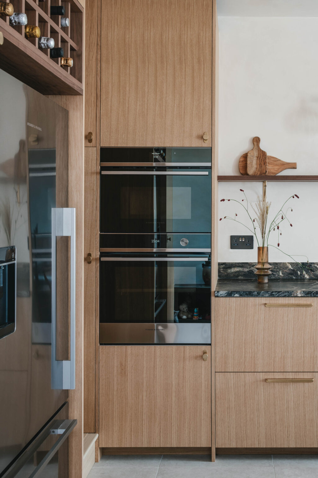 A bright and modern oak wooden kitchen with a built in black oven and black marble worktops. There is a wooden shelf about the worktops with small wooden chopping boards on. There is a silver fridge in the corner of the frame.