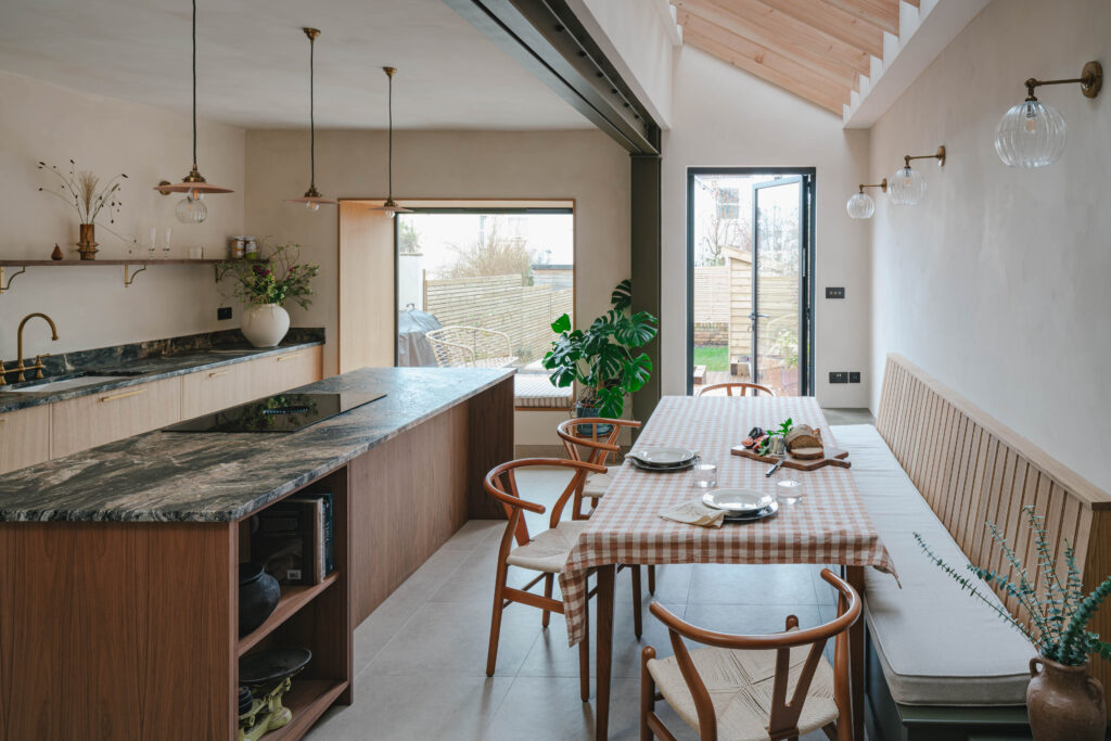 Open-plan kitchen and dining space with a stone-topped wooden island and induction hob beside a long dining table with a checkered tablecloth, wooden chairs, and a built-in bench, lit by pendant and wall lights, with indoor plants and glass doors opening to a garden beneath a sloped timber ceiling.