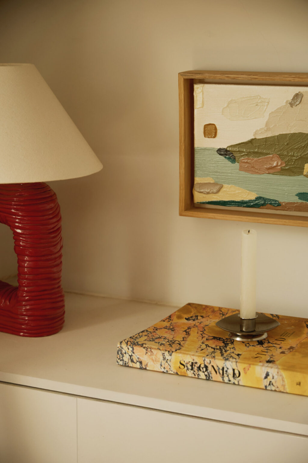 Close-up of home styling featuring a bright red lamp base, book, candle and landscape painting by Rosa Roig-Fiol