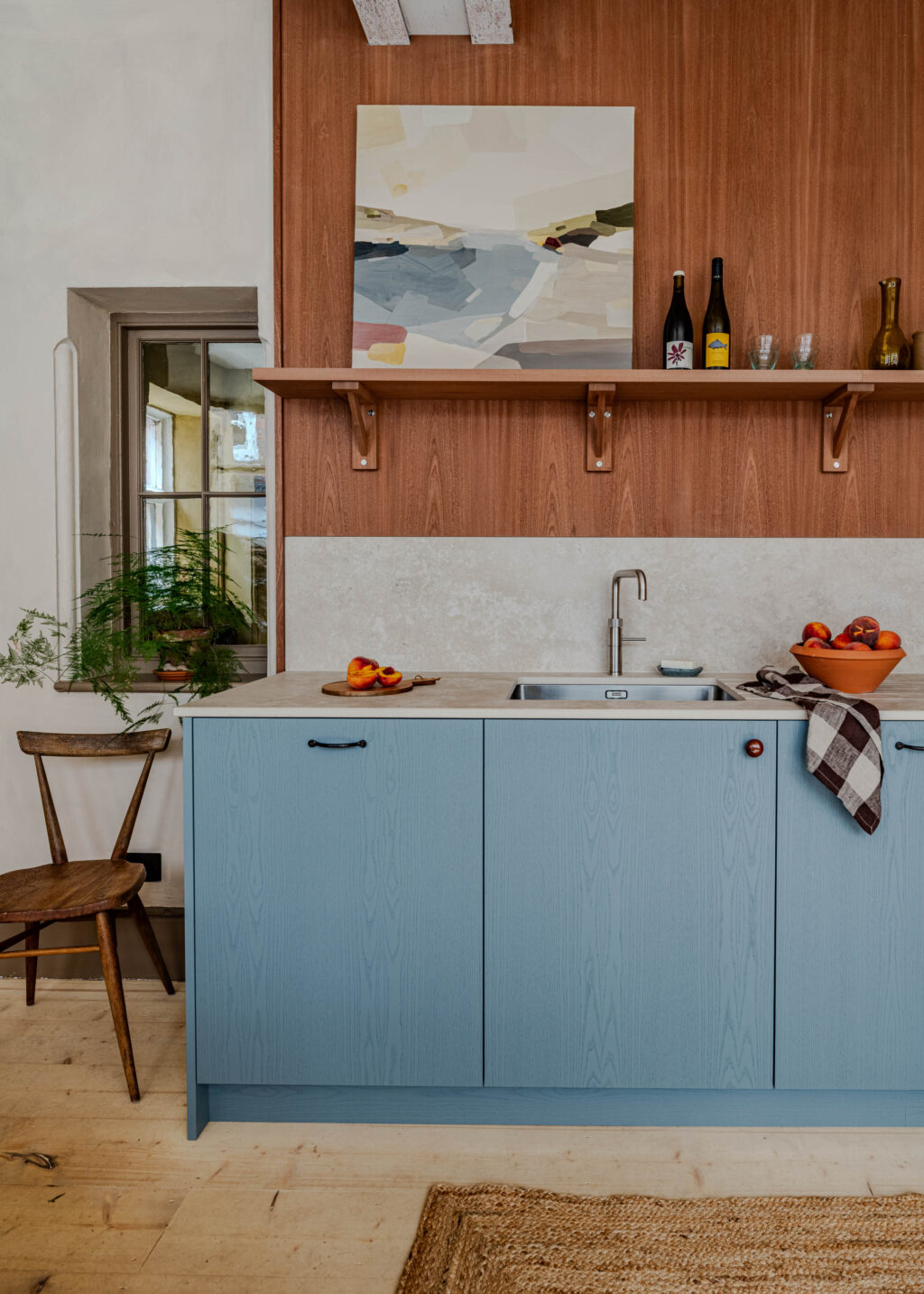 Kitchen interior with blue cabinetry, timber wall and open shelf, viewed from the side with wooden chair.