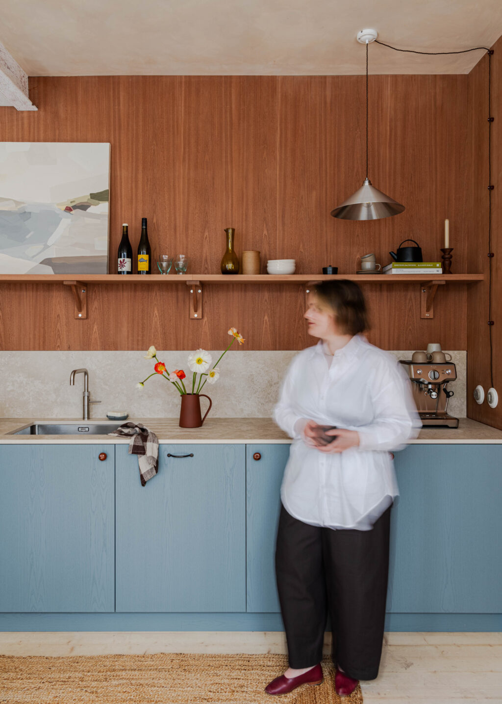 Woman moving through a small kitchen with blue cabinetry, timber wall panelling, open shelving and espresso machine.