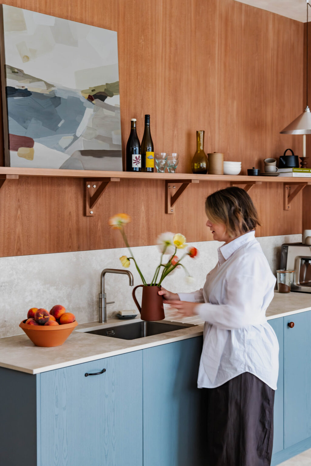 Woman arranging flowers in a terracotta jug beside sink in a timber-lined kitchen with blue cabinets.