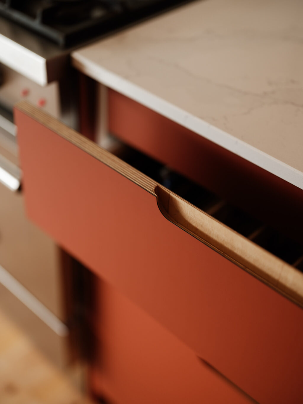 Angled close-up of an open drawer showing layered plywood edges and a carved wooden pull on a red cabinet front.