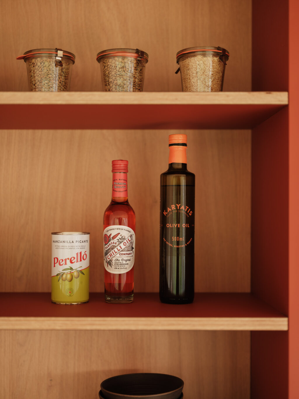 Open shelving in a terracotta cabinet displaying jars of grains, bottles, ceramics, and stacked books against a wood interior.