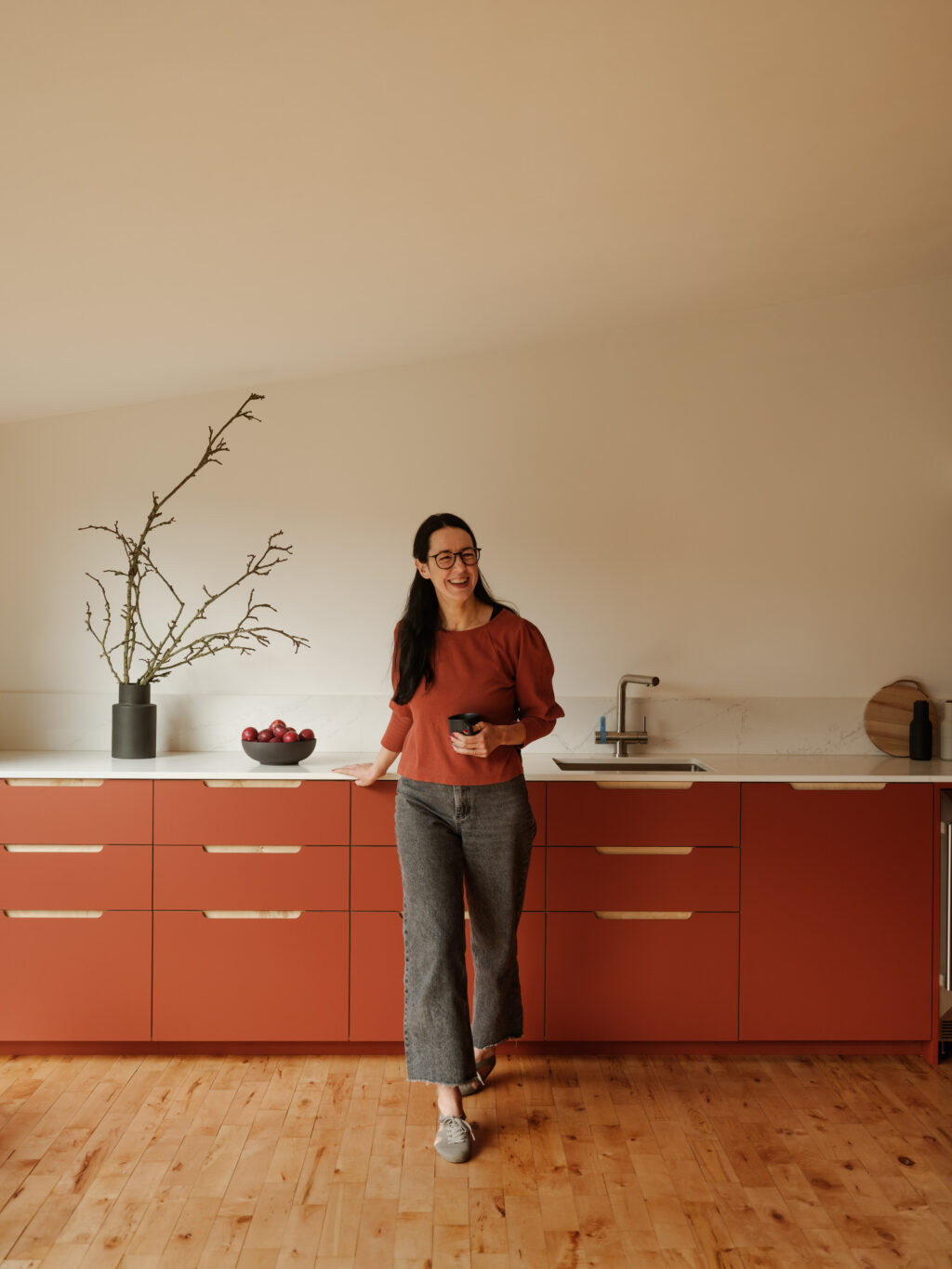 Woman standing in kitchen holding a mug, smiling in front of terracotta cabinetry and white worktop.