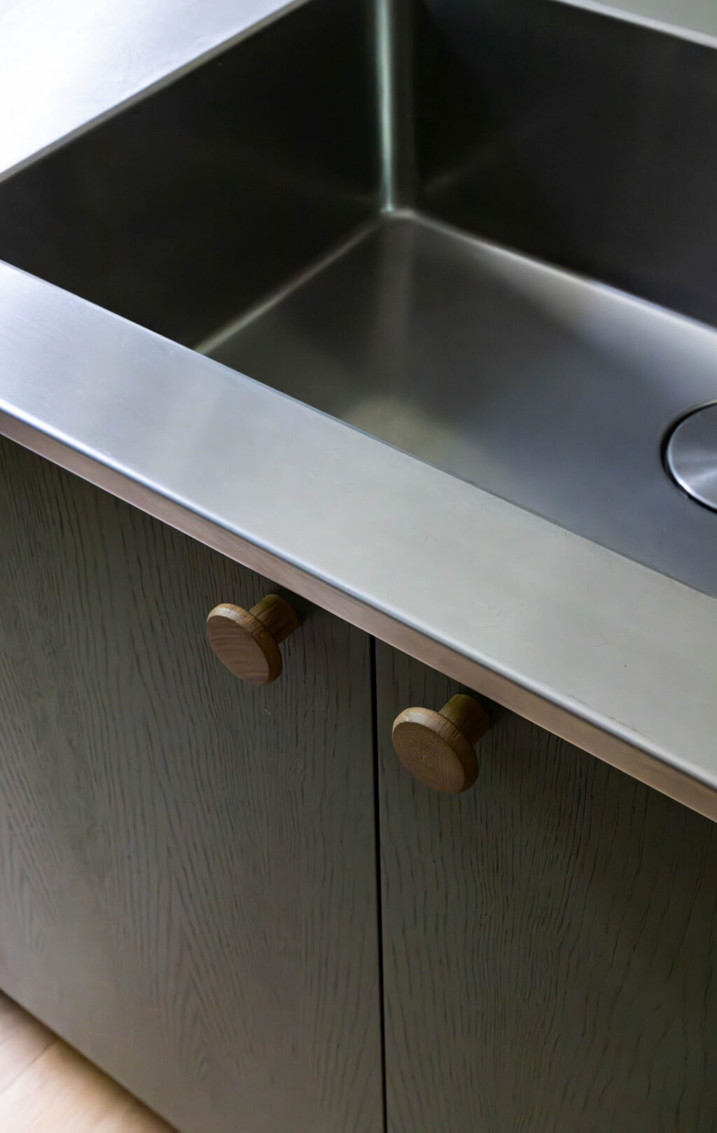 Modern ash veneer kitchen cabinets with wooden handles beneath stainless steel worktop and sink, handcrafted cabinetry detail