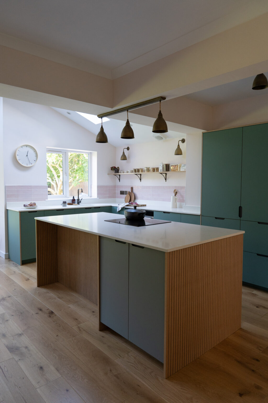 Wide angled view of the full kitchen with central island, green cabinets, wooden flooring, window light, and soft pink tiled backsplash.