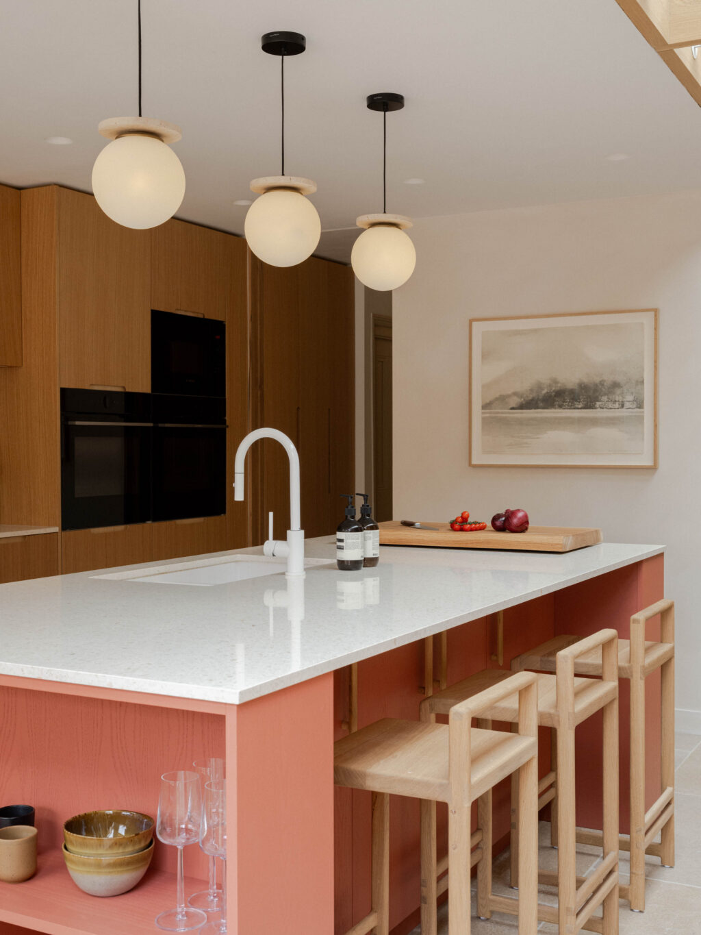 HUSK x Another Country Devon Red Fallow kitchen island with light terrazzo worktop, solid oak pull-bar handles and oak bar stools beneath globe pendant lighting
