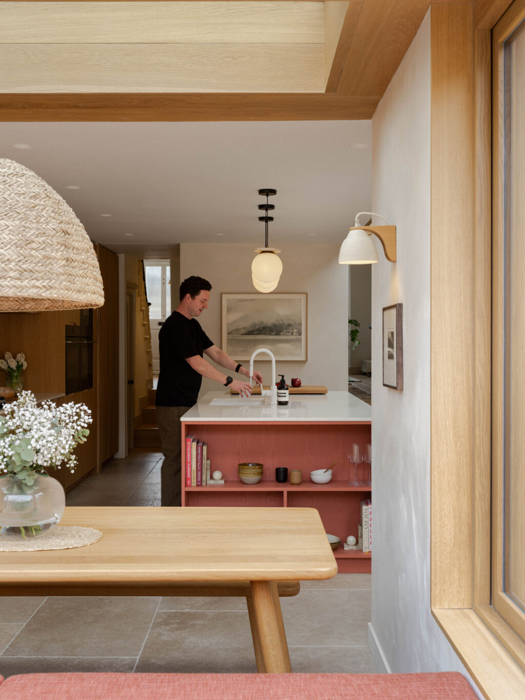 Man at sink on kitchen island in Devon Red Fallow finish with white terrazzo worktop positioned between the dining area and living space beneath oak ceiling beams