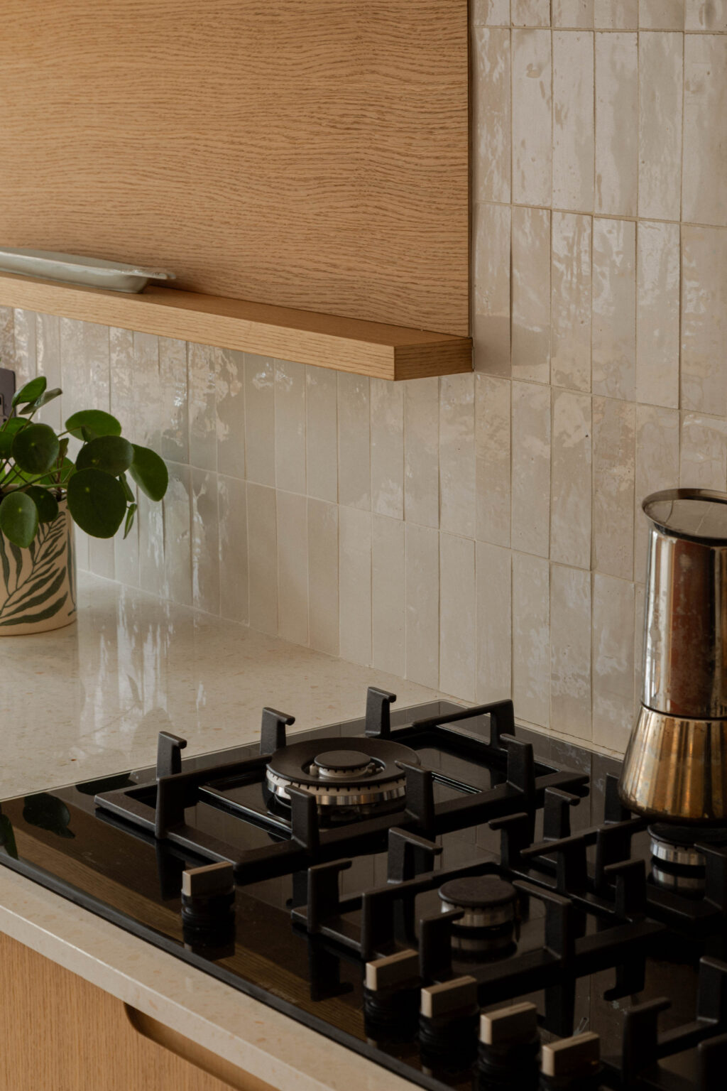 Detail of glossy cream zellige tile splashback with integrated gas hob, terrazzo worktop and Natural Oak Veneer custom shelving and cabinetry beneath