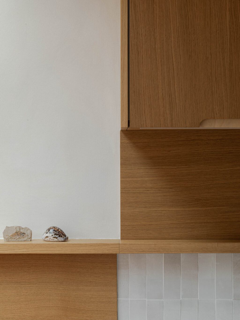 Detail of integrated oak shelving and cabinetry junction against a cream zellige tile splashback with styled shell objects