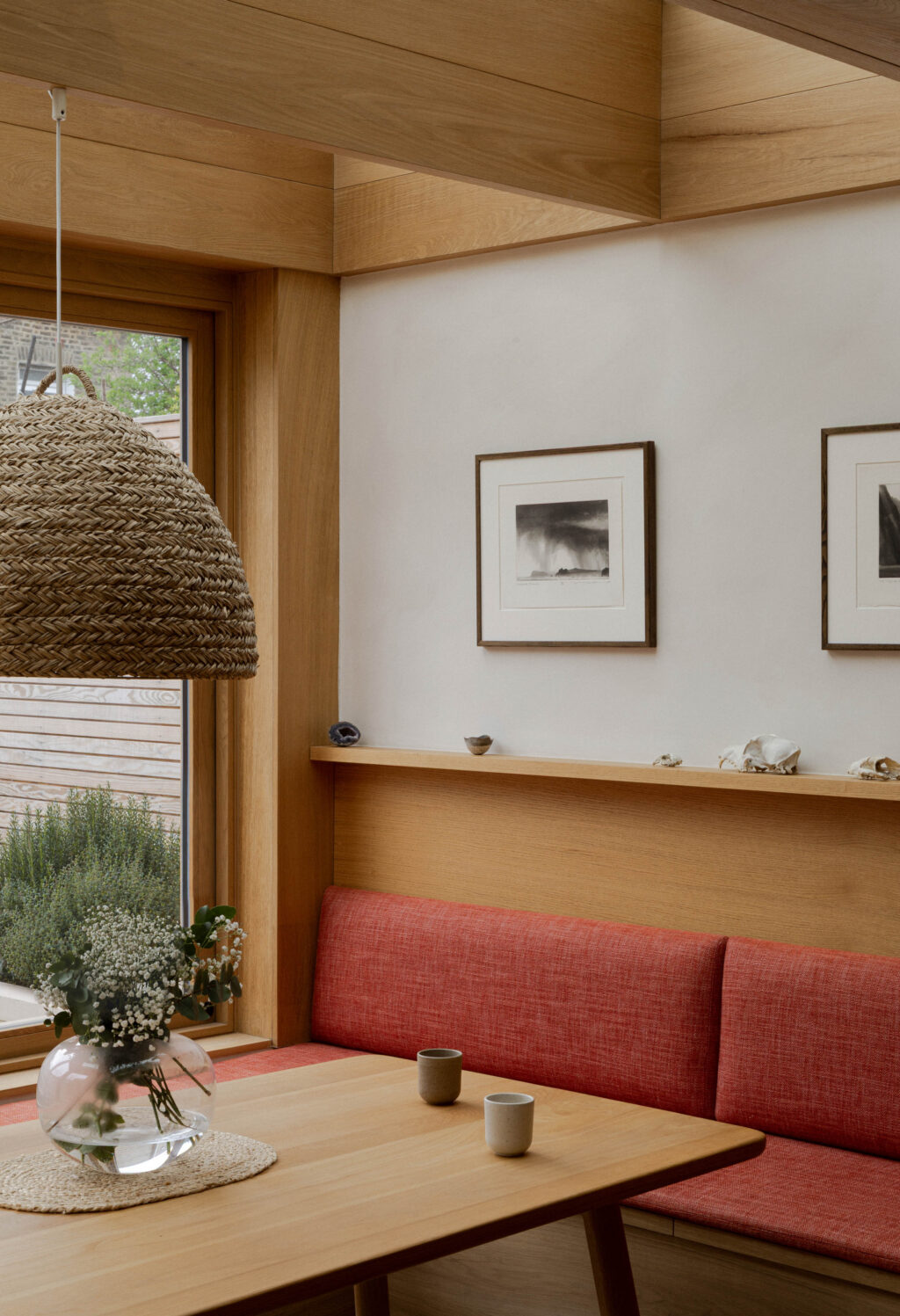 Built-in coral banquette seating beneath oak shelving with woven pendant lighting and garden views