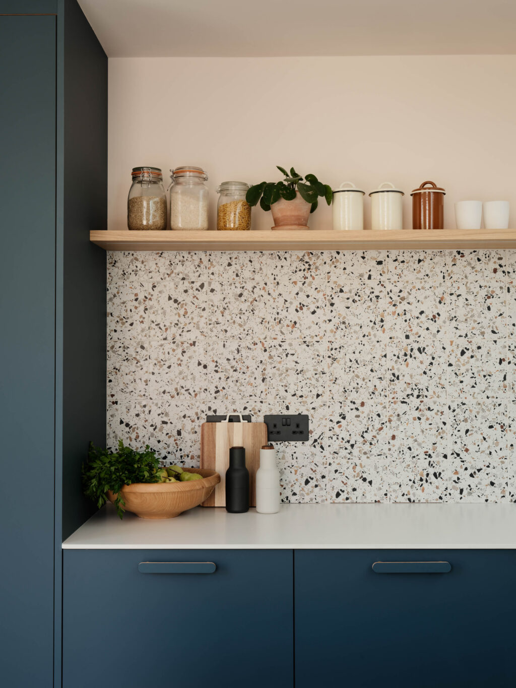 Terrazzo-effect tiled splashback with a natural oak floating shelf, styled with storage jars, houseplants and kitchen accessories.
