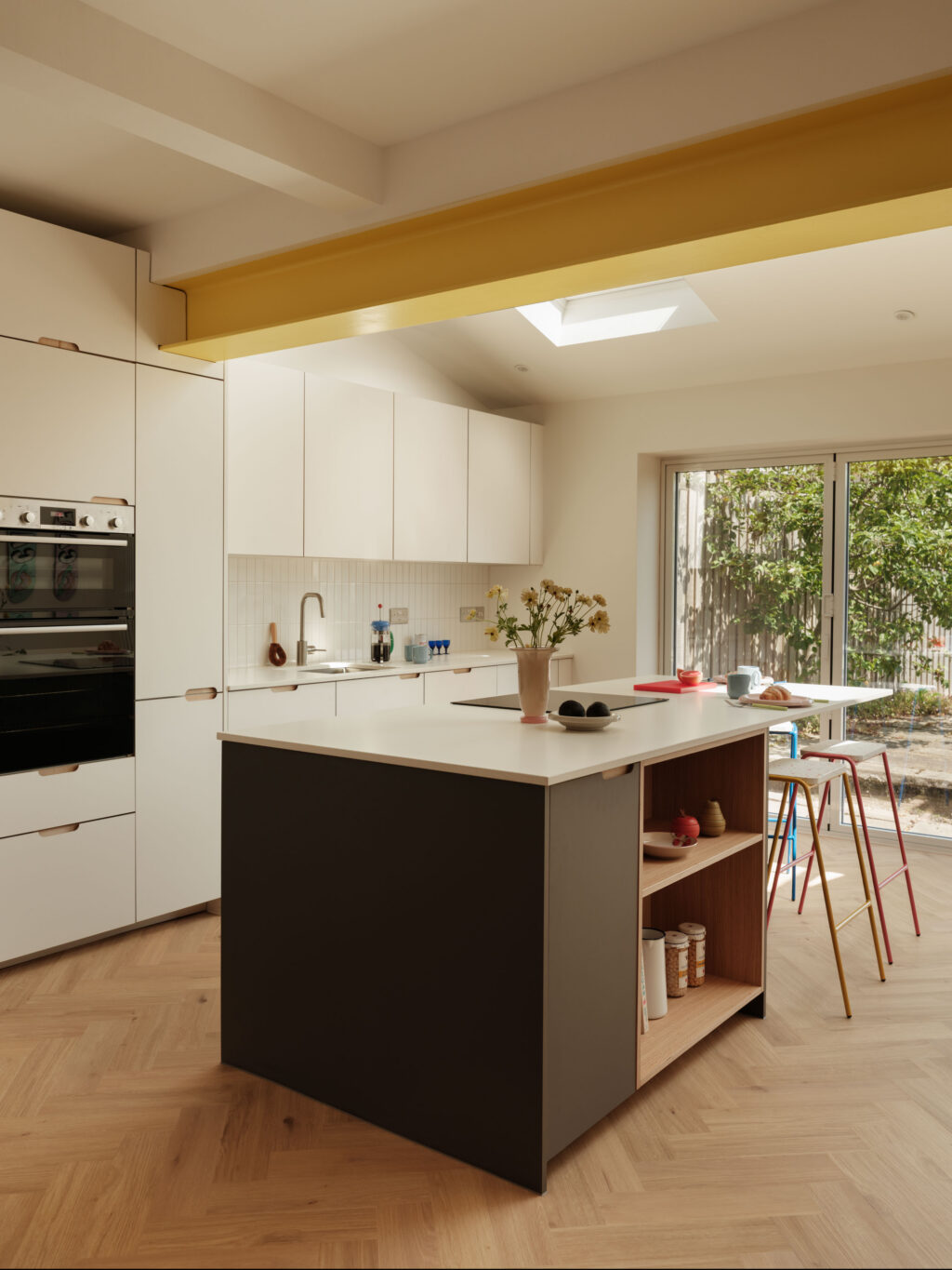 Kitchen island beneath a bright yellow RSJ beam featuring FENIX Bianco Male cabinetry, FENIX Grigio Aragona island panels and natural oak open shelving.