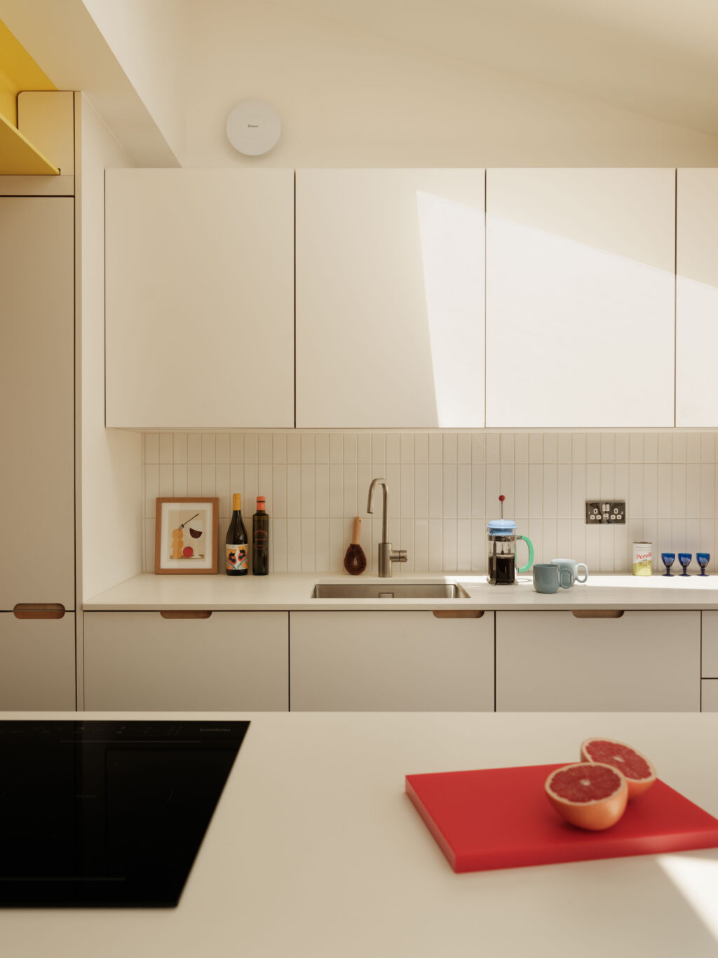 FENIX Bianco Male cabinetry and sink run with exposed ply scallop handles, vertical tiled splashback and Neolith Just White worktops illuminated by natural light.