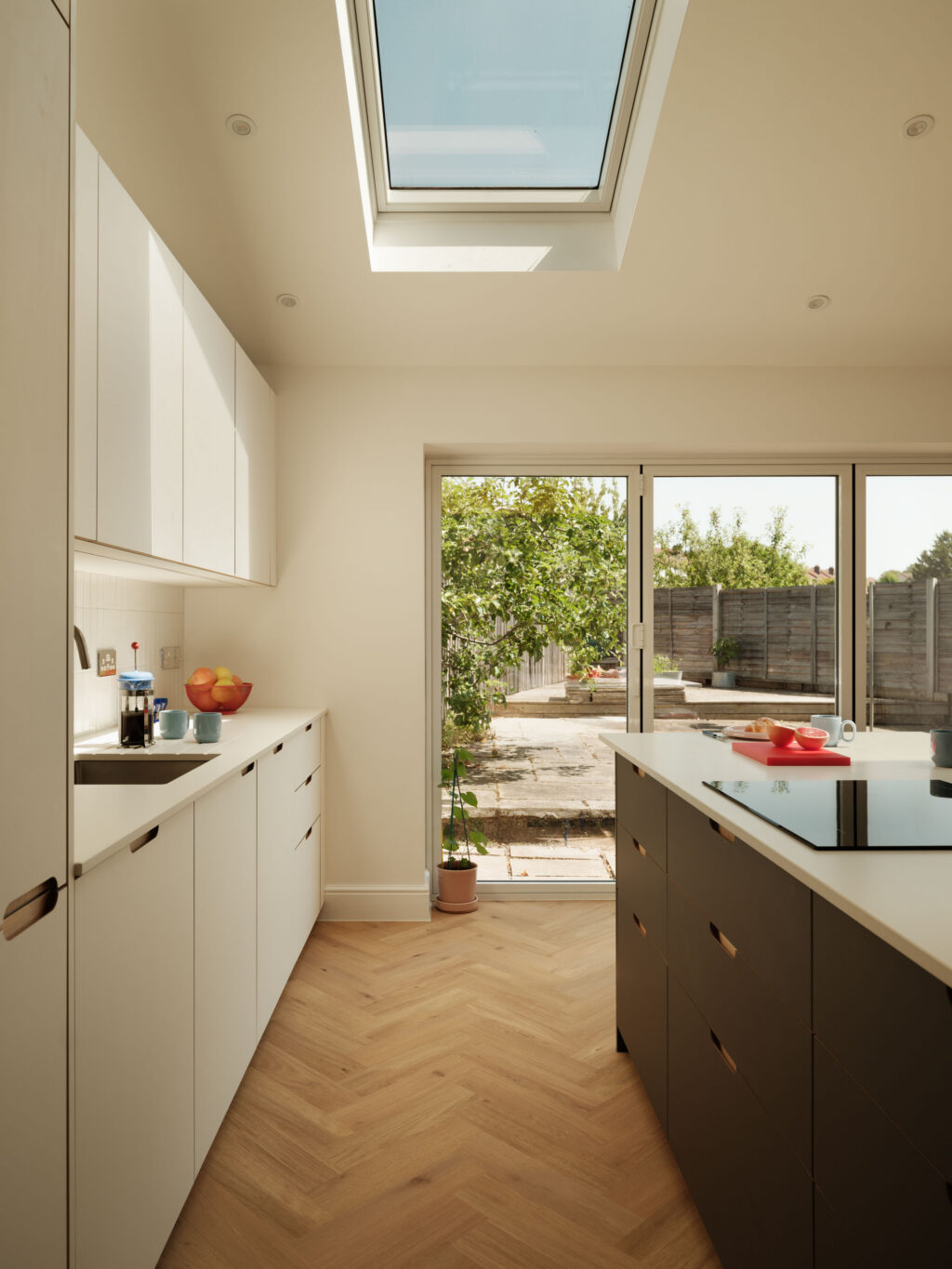 View through The St George Kitchen towards the garden, featuring a large rooflight, FENIX Bianco Male cabinetry, FENIX Grigio Aragona island with induction hob and Neolith Just White worktops.