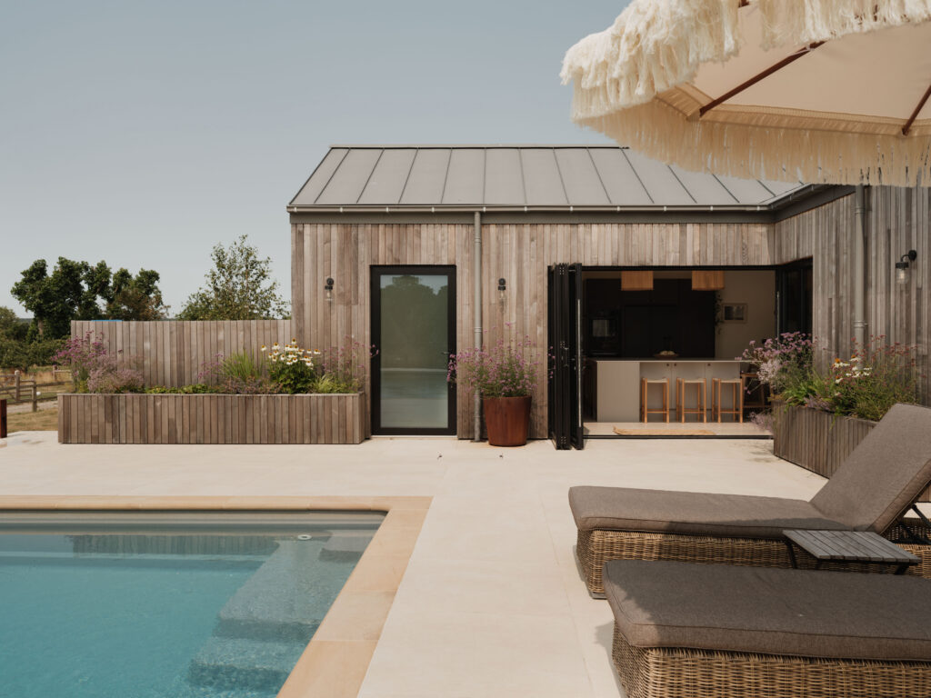 Contemporary timber-clad home with fully open bifold doors connecting the HUSK kitchen to a landscaped terrace and outdoor swimming pool.