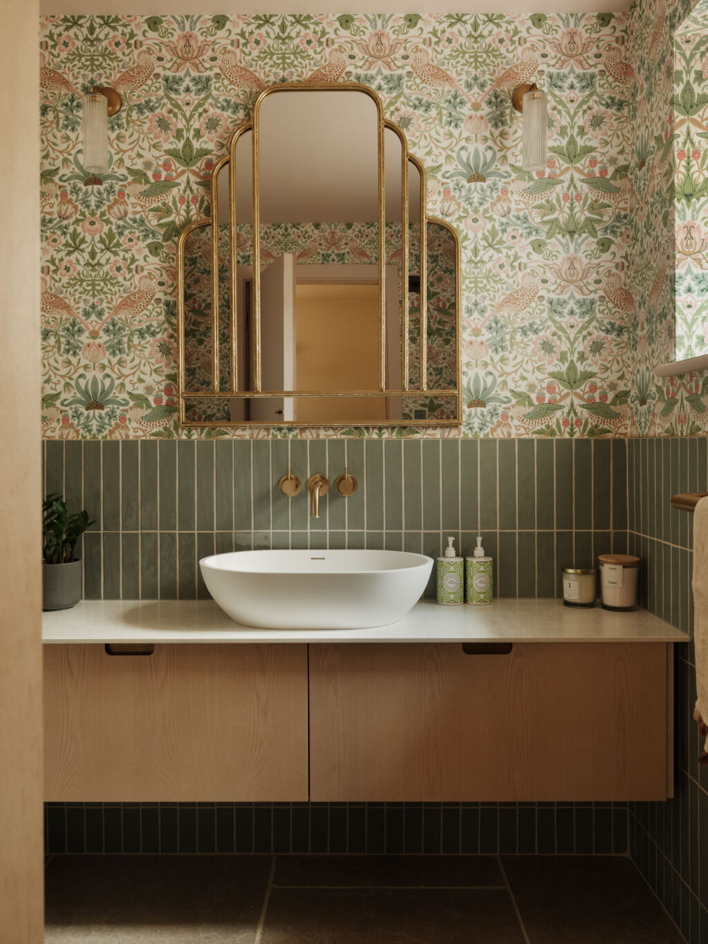 White Tint Ash on Ply bathroom vanity with integrated scallop handles and custom brass inlay beneath a sculptural brass-framed mirror, complemented by green vertical tiles and botanical wallpaper.