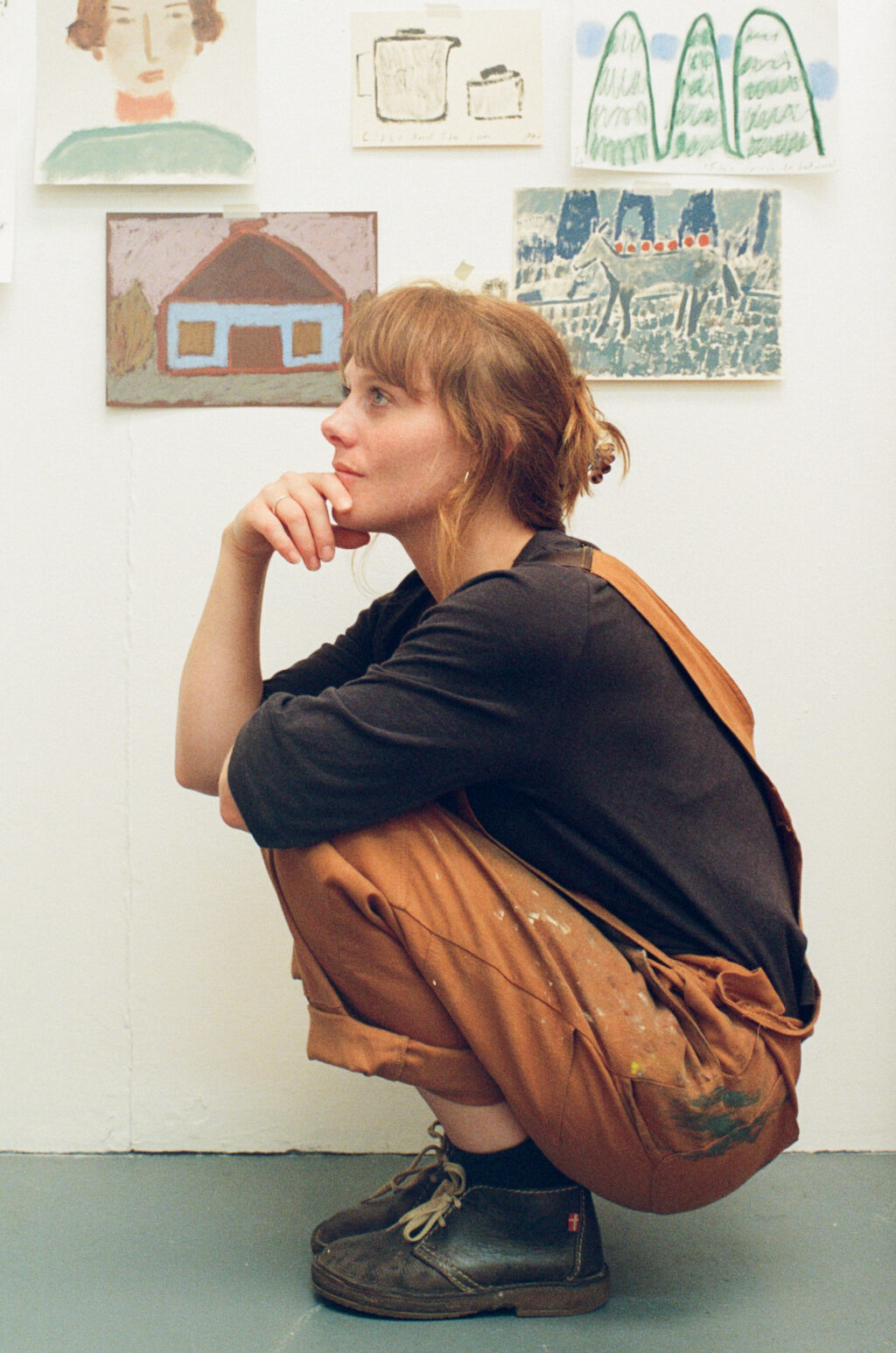 Artist Jay Harper crouching in her Bristol studio, wearing paint-splattered overalls with paintings displayed behind her.