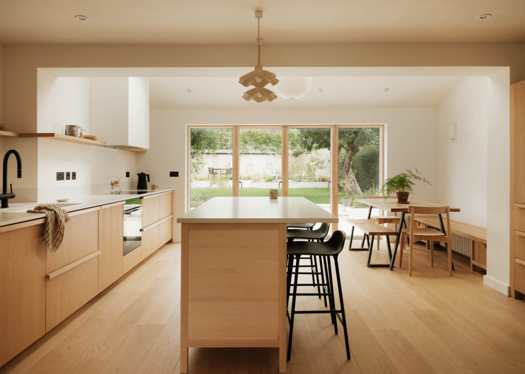 Wide view of the open-plan Beckington Kitchen with Natural Oak cabinetry, central island, dining area, built-in bench seating and garden doors.