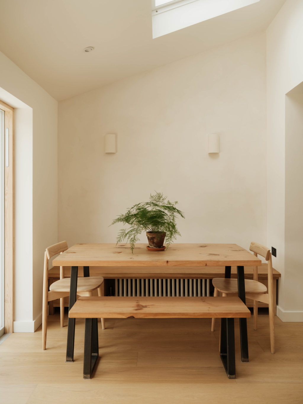 Dining area in The Beckington Kitchen with a timber table and bench, wooden chairs, built-in bench seating and a potted fern beneath the skylight.