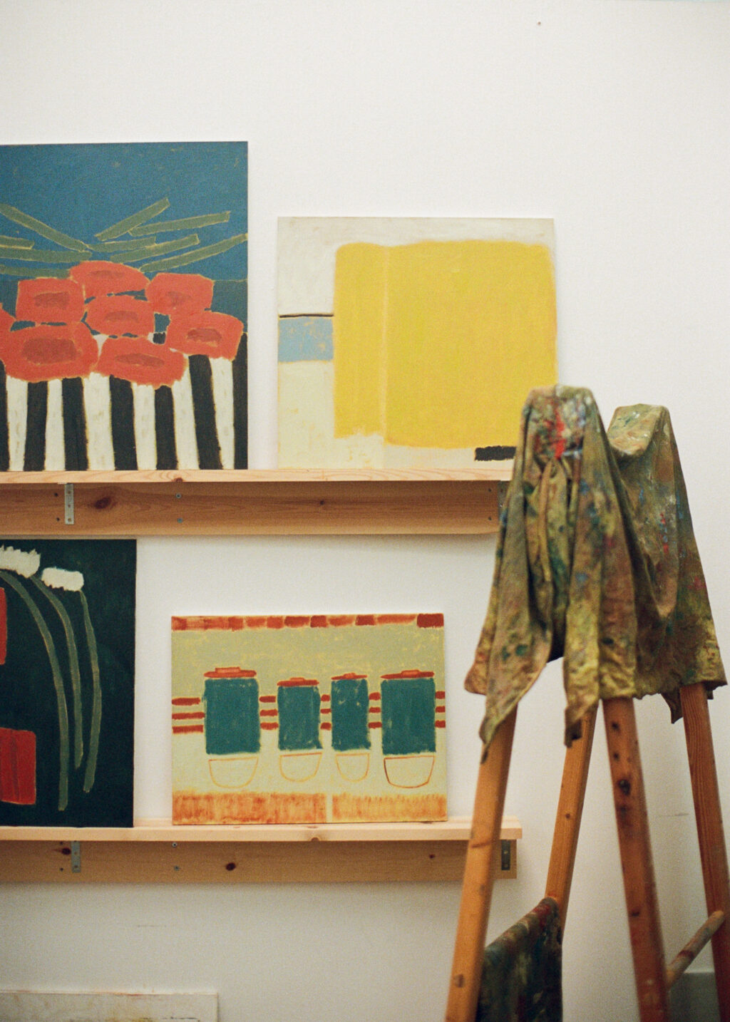 Colourful paintings displayed on wooden shelves in Jay Harper’s studio, with paint-splattered overalls draped over a stool.